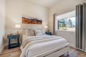 A bedroom with a bed, two bedside tables, a nightstand, a window with curtains, and a painting on the wall.