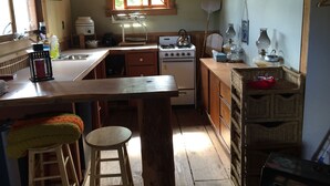A compact kitchen with a central island, wooden cabinets, and a window providing natural light. - Cozy Cabin on a small Off Grid Homestead (Shawnigan Lake)