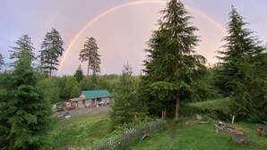 A house with a green roof surrounded by dense forest and a rainbow in the background. - Cozy Cabin on a small Off Grid Homestead (Shawnigan Lake)