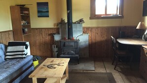 A cozy living room with a sofa, a wooden table, a fireplace, and a window. - Cozy Cabin on a small Off Grid Homestead (Shawnigan Lake)