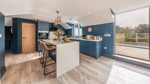 A modern kitchen with a central island, bar stools, and a large window offering a view of the outdoors. - May Cottage Titchwell, Norfolk (Titchwell)