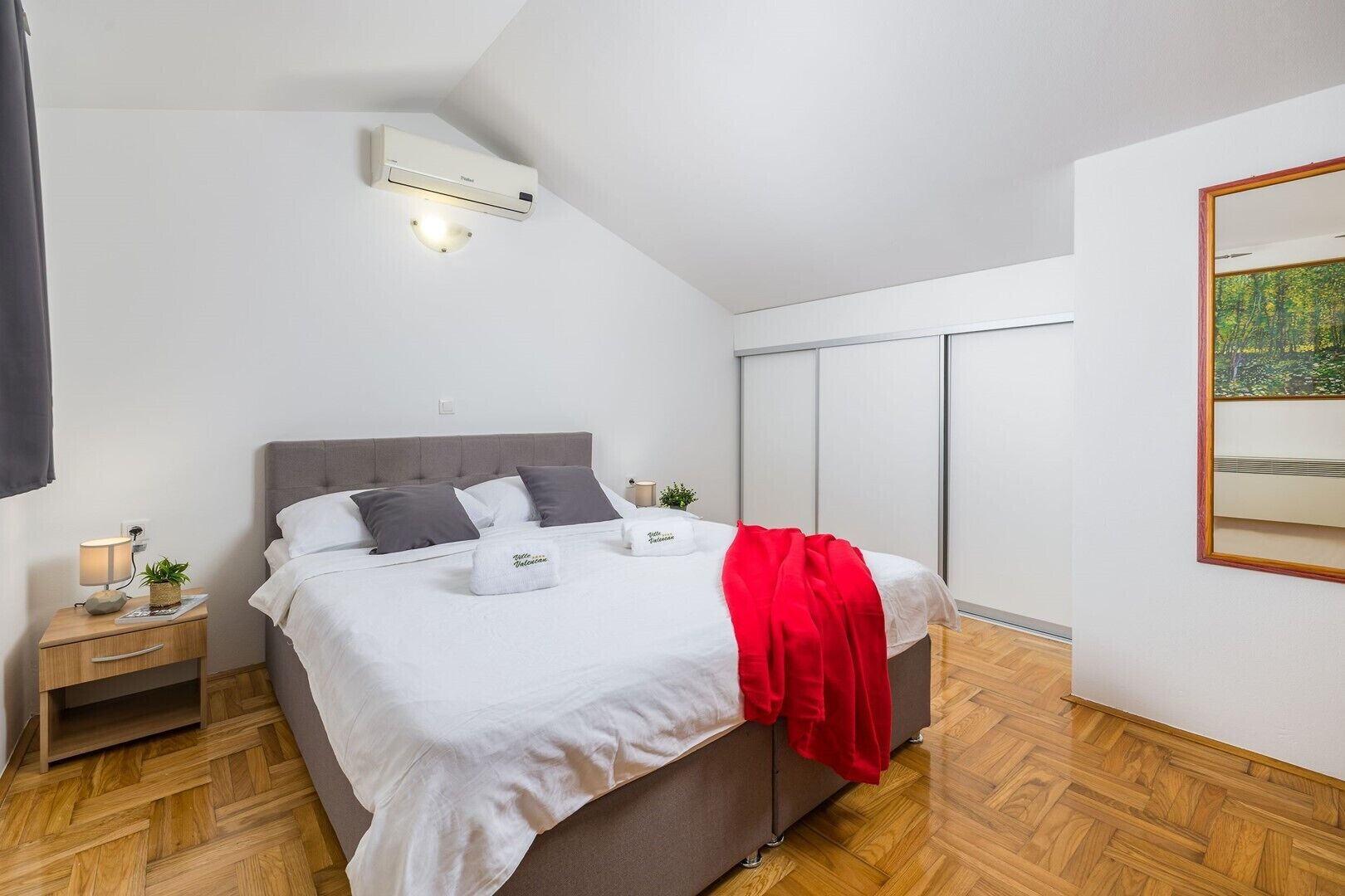 A modern bedroom with a bed, bedside tables, a nightstand, an air conditioning unit, a red throw blanket, and a large mirror.