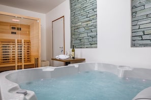 Een spa kamer met een ruime hottub, houten banken, een stenen muur en een kleine tafel met een fles en glazen.