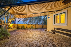 Un patio de piedra con una pérgola de madera, un banco y un edificio con pared amarilla y ventana de marco verde.
