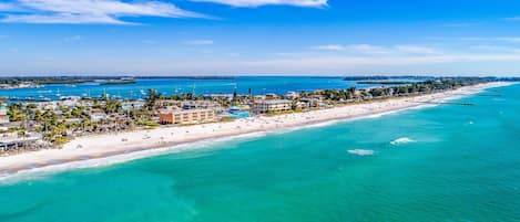 An aerial view of a coastal resort with a long beachfront, clear turquoise waters, and a variety of buildings including hotels and villas.