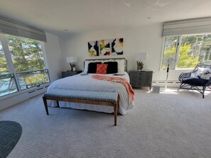 A spacious bedroom with a large bed, a nightstand, a chair, and a window with a view of trees.