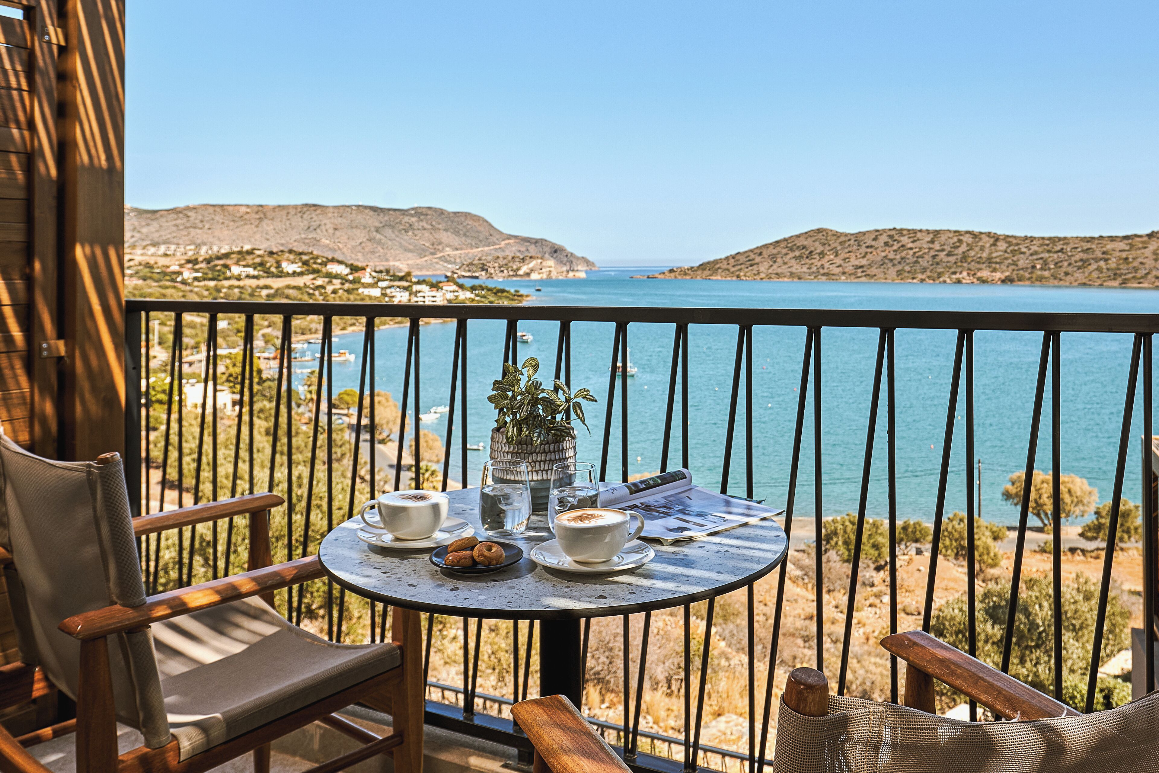 A balcony with a table set for two, overlooking a coastal landscape with hills and a clear sky.