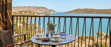 A balcony with a table set for two, overlooking a coastal landscape with hills and a clear sky.