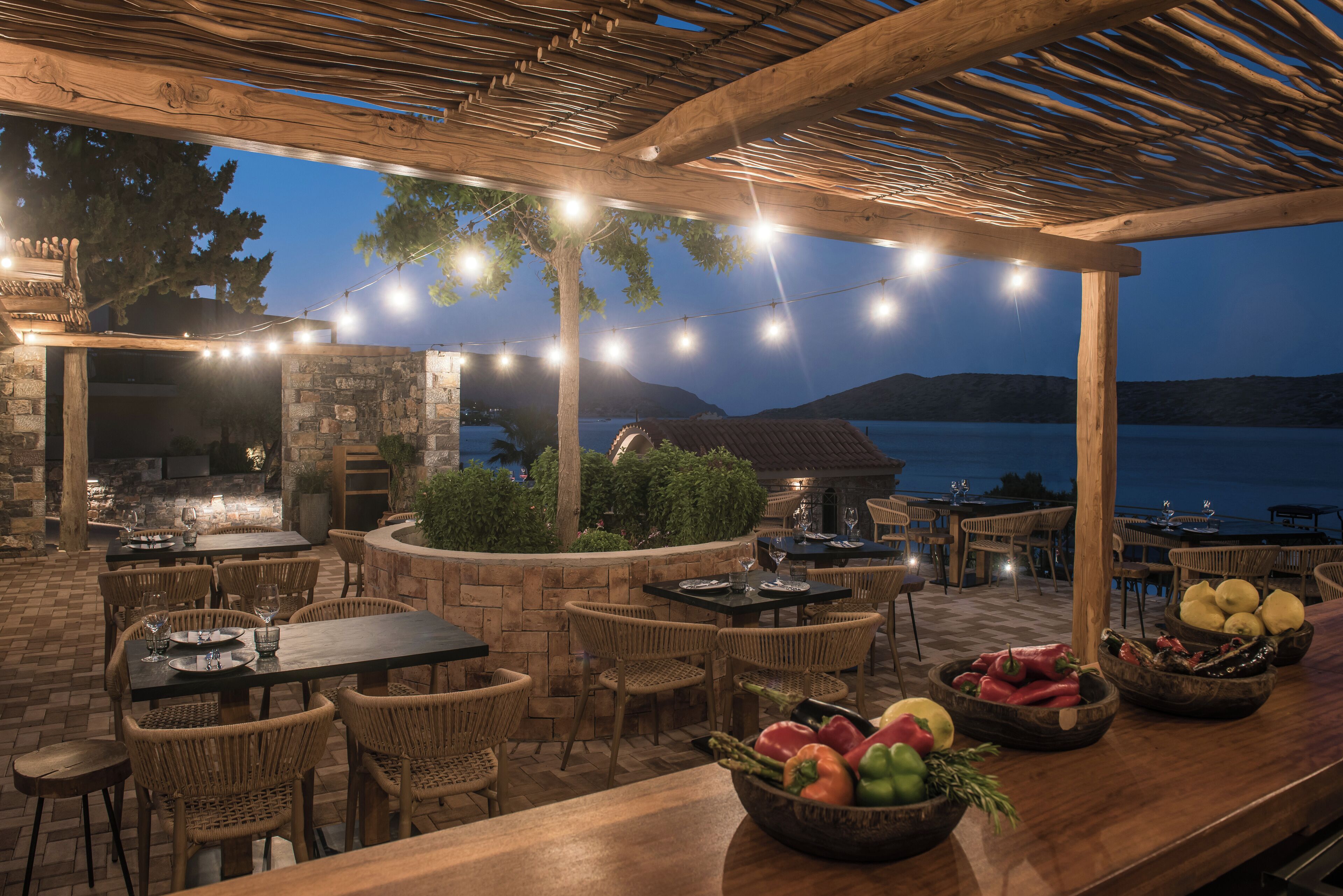 Un área de comedor al aire libre con muros de piedra, pérgola de madera, luces colgantes y vistas al agua.