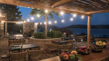 Un área de comedor al aire libre con muros de piedra, pérgola de madera, luces colgantes y vistas al agua.