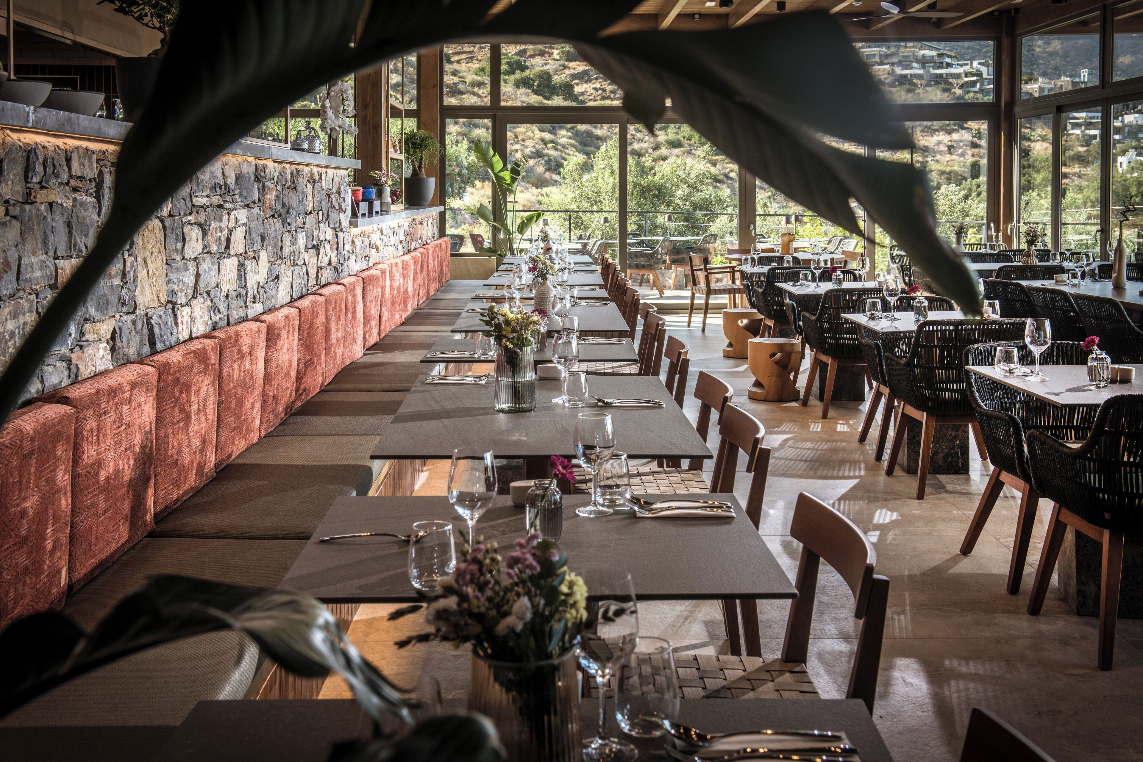 Um restaurante com uma mesa comprida preparada para refeições, uma parede de pedra de um lado e vista para as colinas através de grandes janelas.