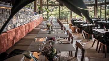 A restaurant with a long table set for dining, a stone wall on one side, and a view of the hills through large windows.