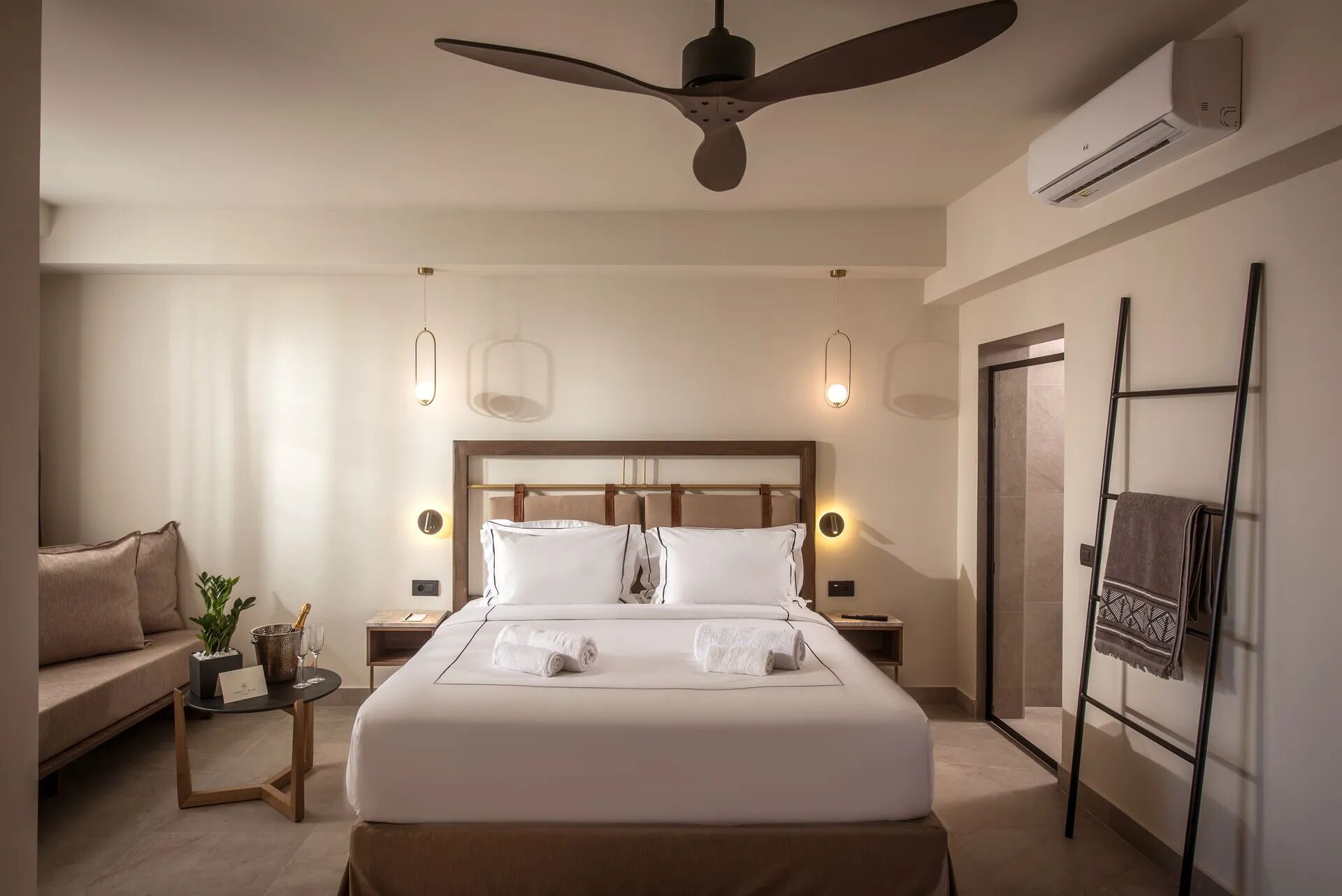 A modern hotel room with a large bed, a ceiling fan, a bedside table with a lamp, a small plant, and a wall-mounted air conditioner.