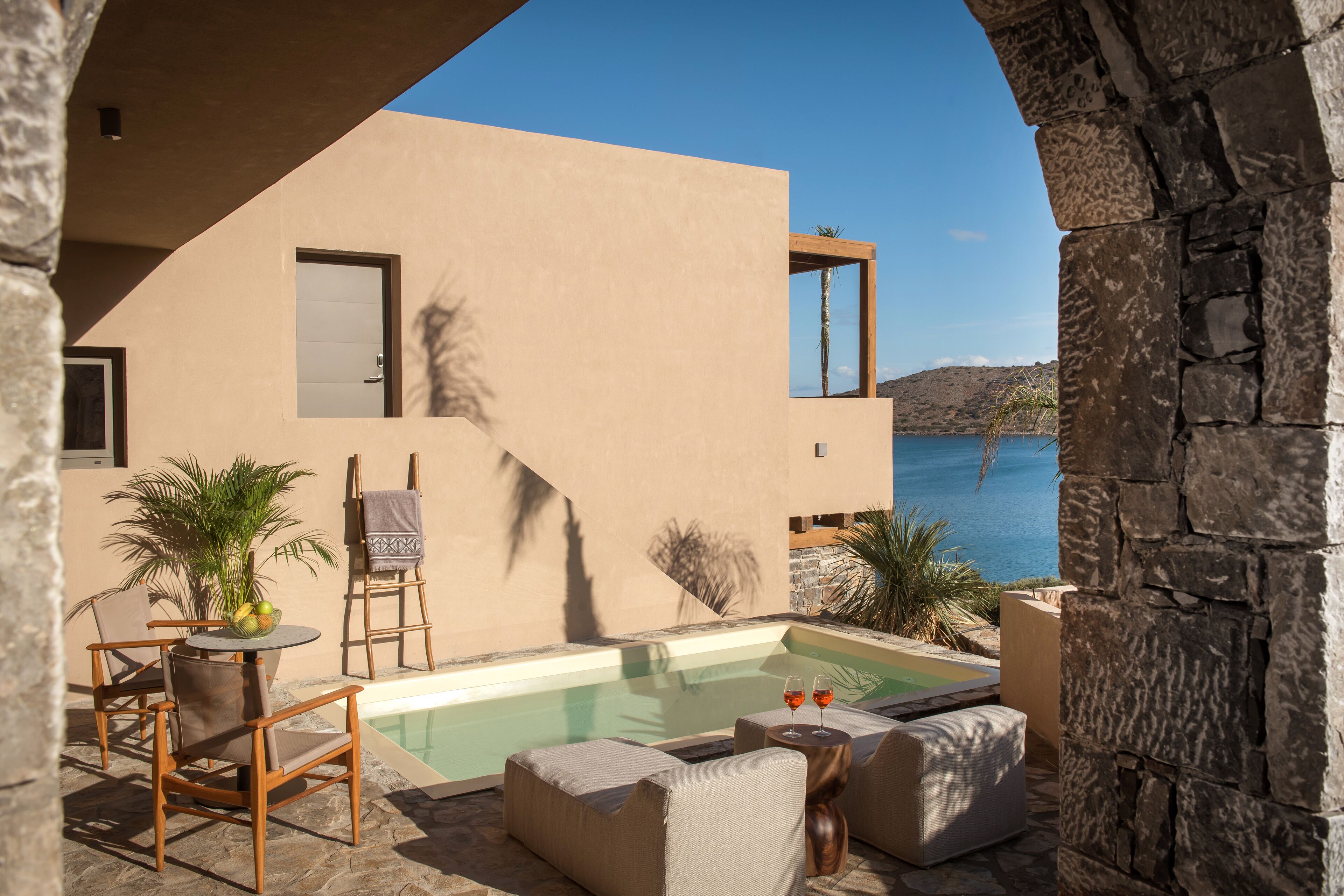 A modern outdoor pool area with a stone wall, a table with chairs, and a view of the sea.