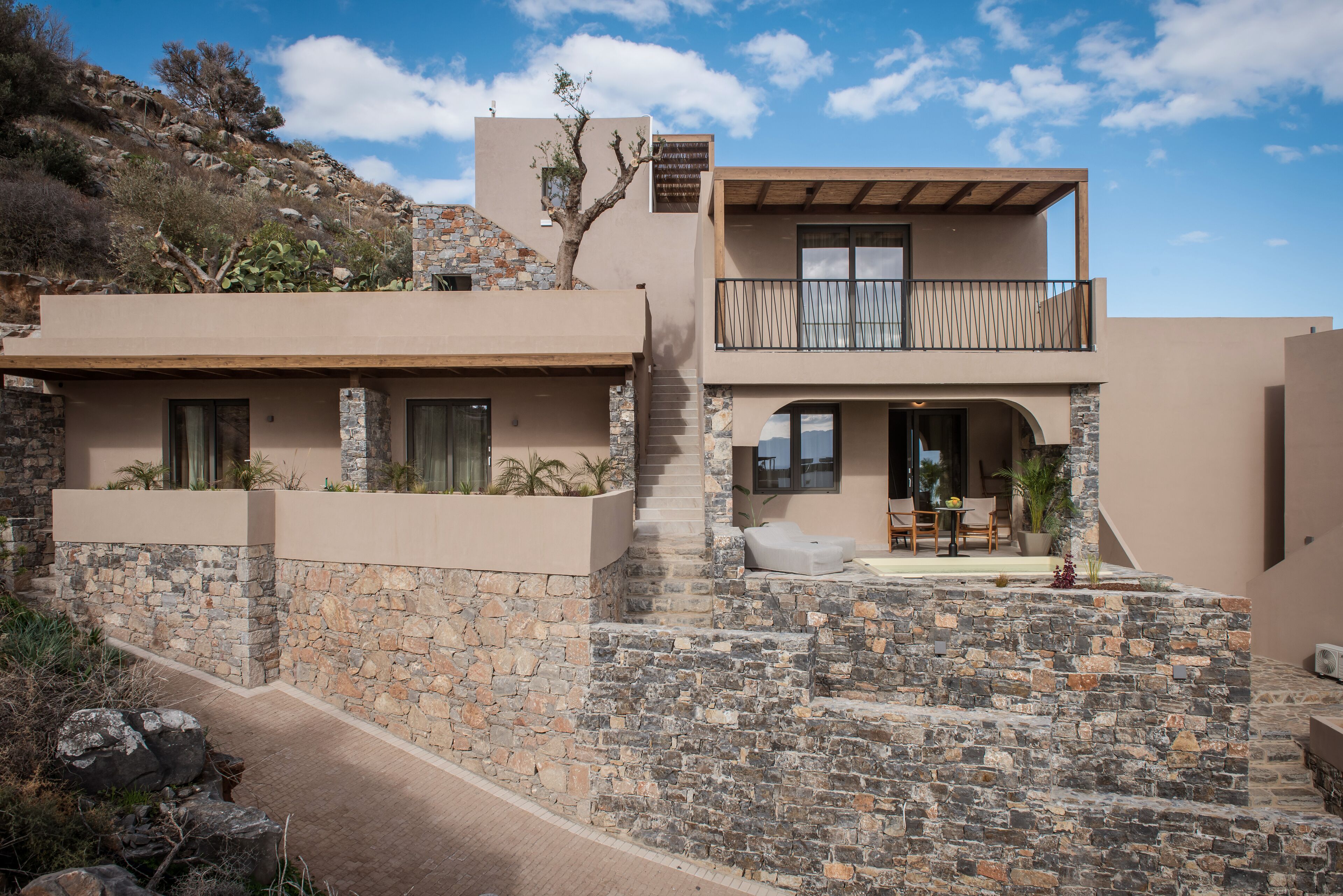 Una casa moderna de dos pisos amb façana de pedra, un balcó i una zona de pati amb mobiliari exterior.