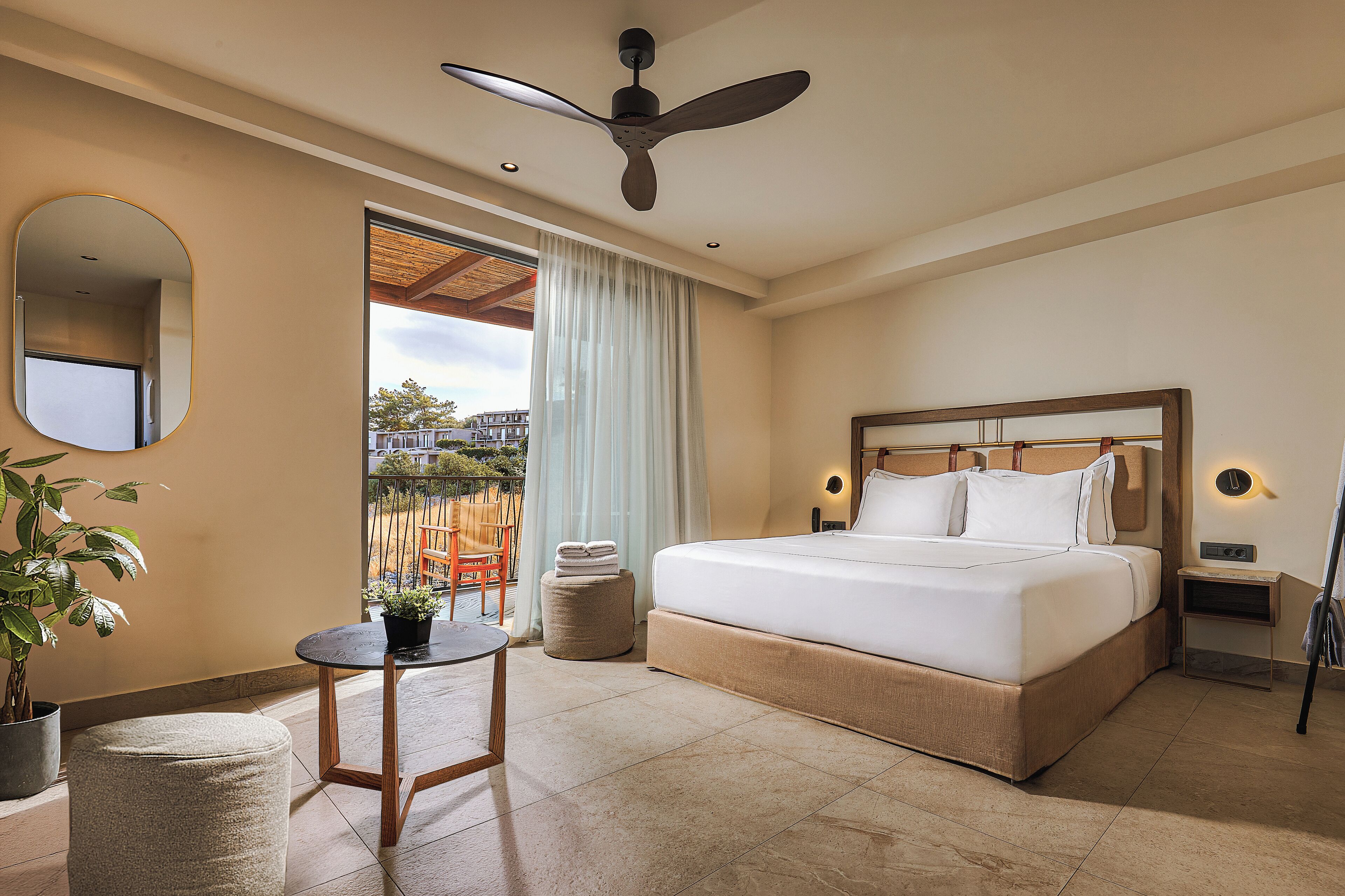 A hotel room with a large bed, a ceiling fan, a small table, a potted plant, and a view of the outside.
