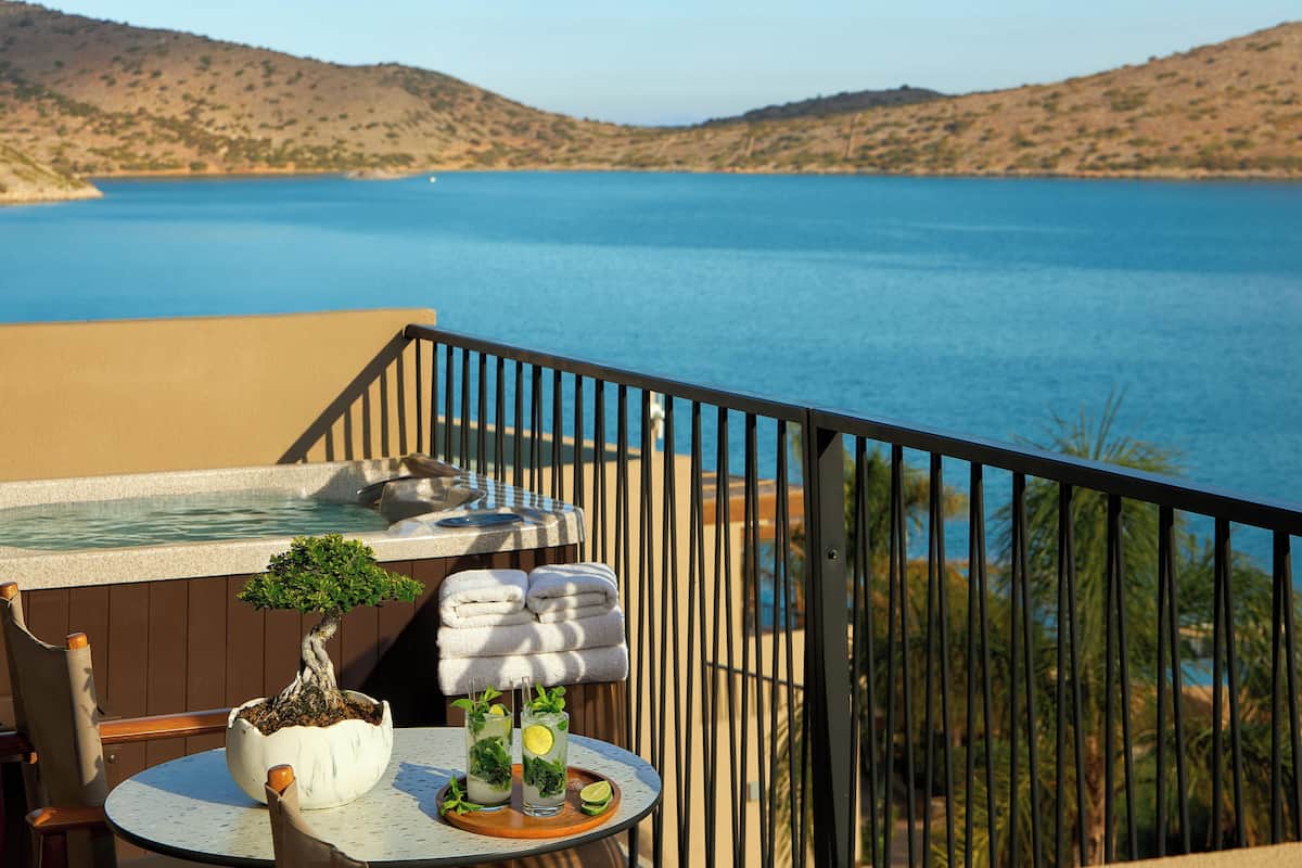 A balcony with a view of a large body of water, a potted bonsai tree, a table with drinks, and towels.