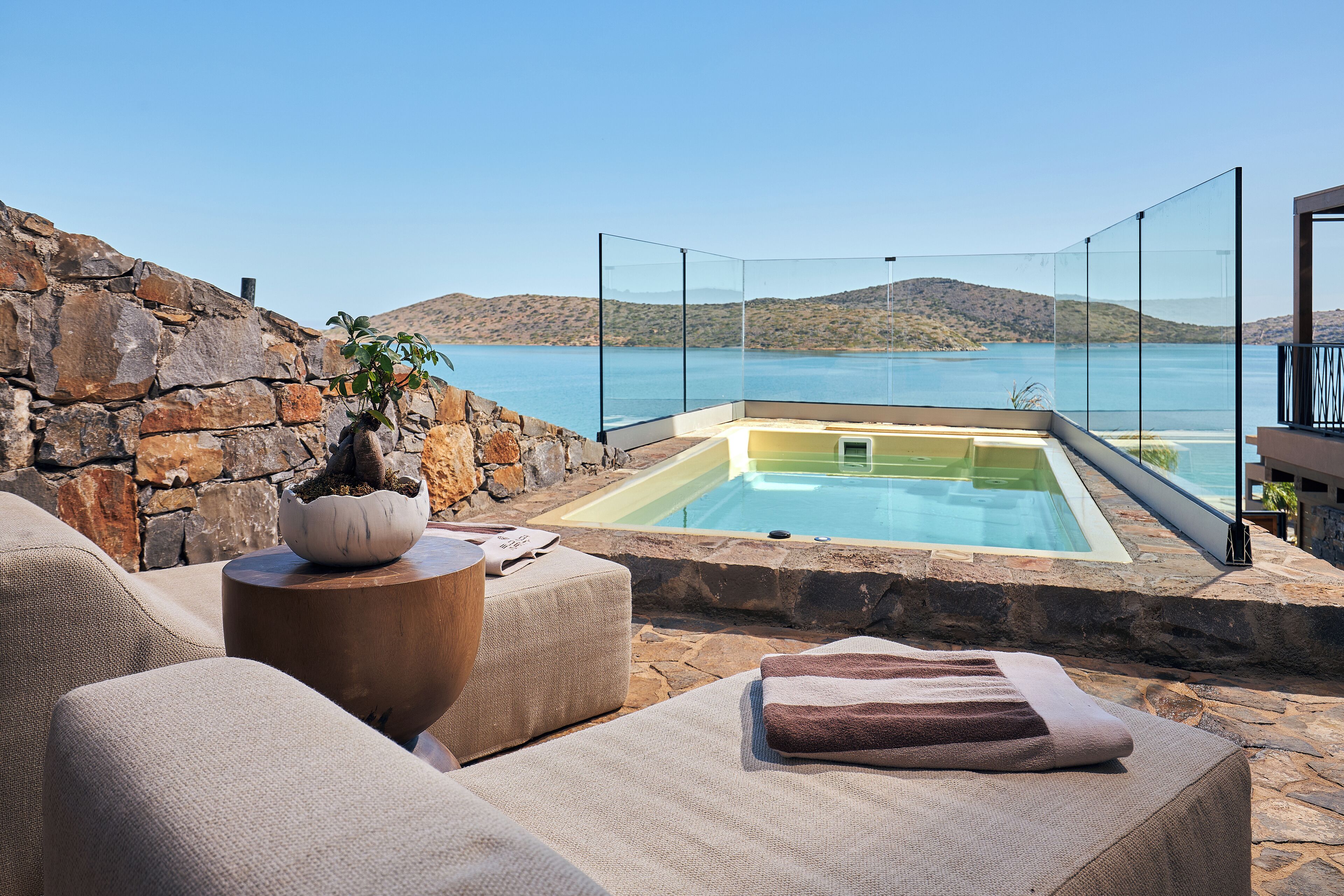 A stone wall, a glass-enclosed infinity pool, and a lounge area with a view of the water.