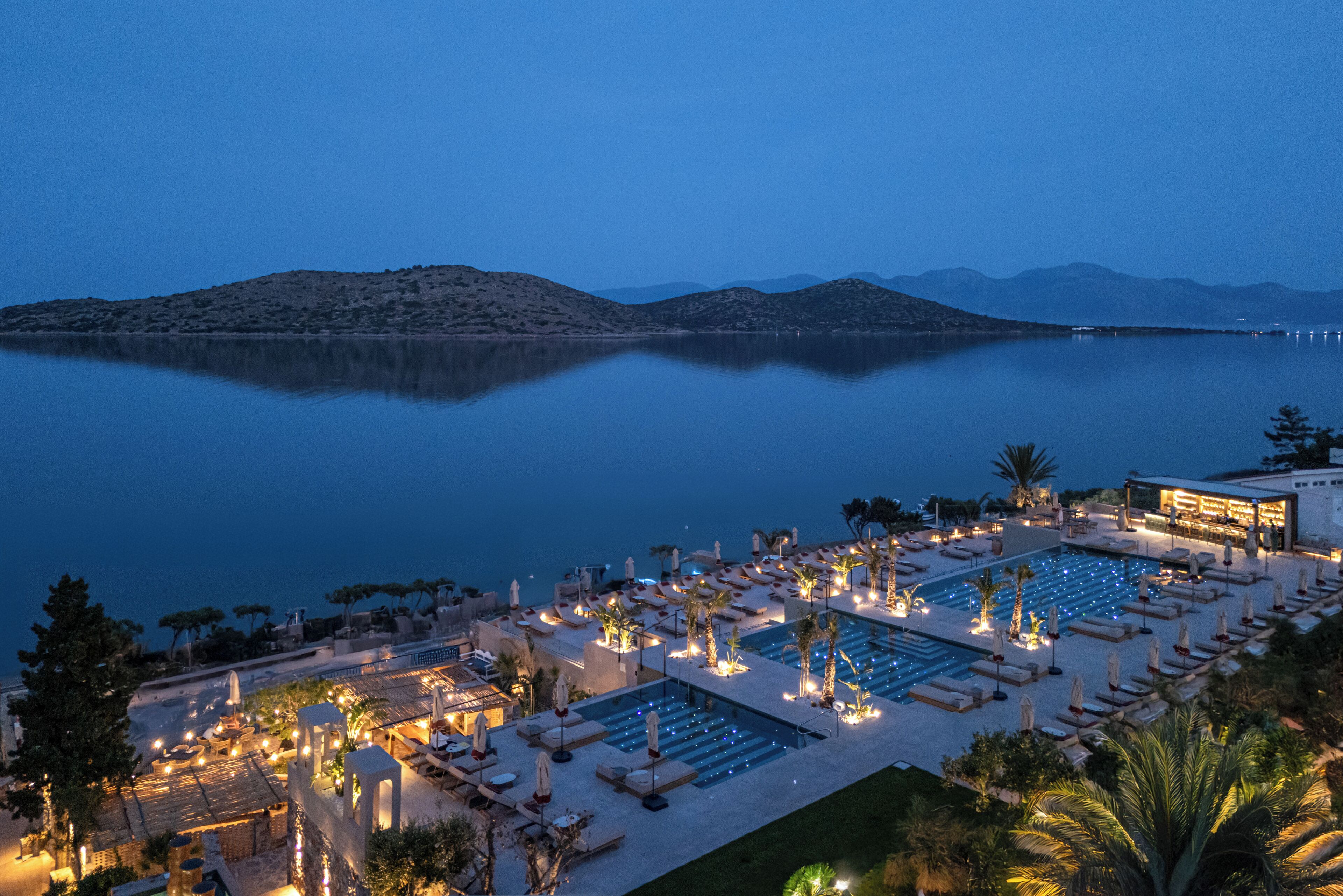 A resort with multiple swimming pools, sun loungers, and a view of a calm lake with mountains in the background.
