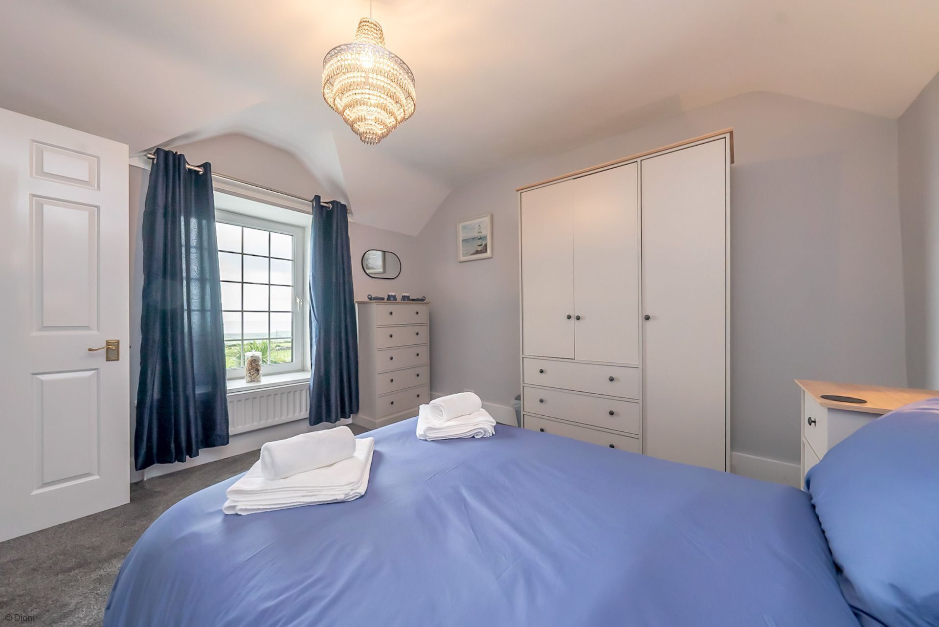 A bedroom with a bed, a wardrobe, a dresser, a window with curtains, and a chandelier.