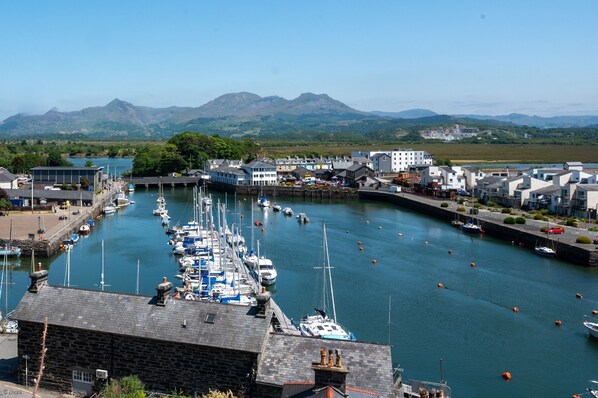 A marina with boats docked, a stone building in the foreground, and a town with buildings and mountains in the background.