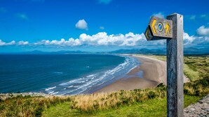 A wooden signpost with a walking symbol and a circular emblem, overlooking a beach with waves and distant mountains under a clear sky.