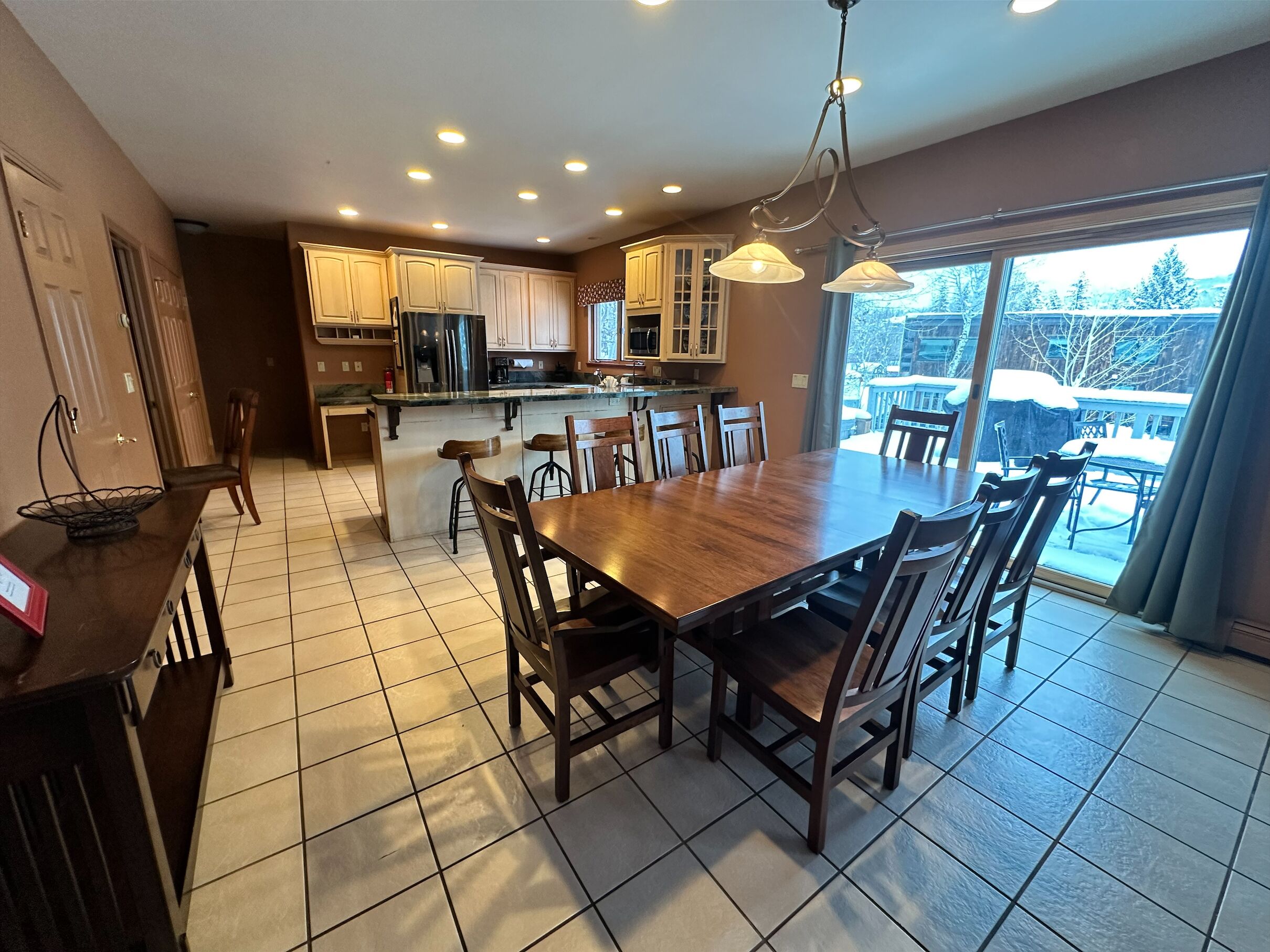 A modern kitchen with a central island, wooden dining table, and chairs, with a view of a snowy backyard through large windows.