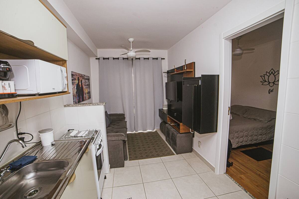 A compact kitchen with a microwave, stove, sink, and a small refrigerator. There is a ceiling fan, a bed with a wooden headboard, and a wall-mounted TV.
