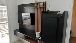 A modern living room with a flat-screen TV mounted on a black wall, a wooden shelf above it, and a black refrigerator below. - Chácara Santo Antonio - Laguna 430 (São Paulo)