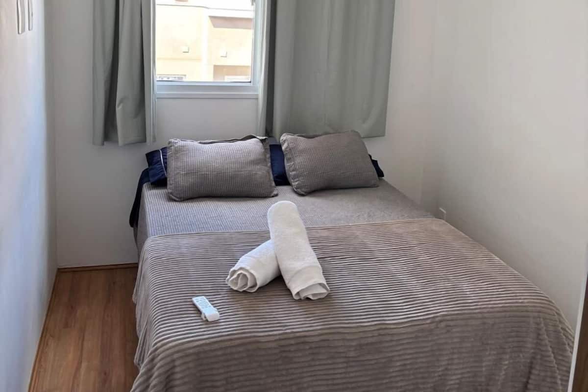 A small, neatly arranged bedroom with a single bed, a window with curtains, a ceiling fan, and a view of buildings outside.