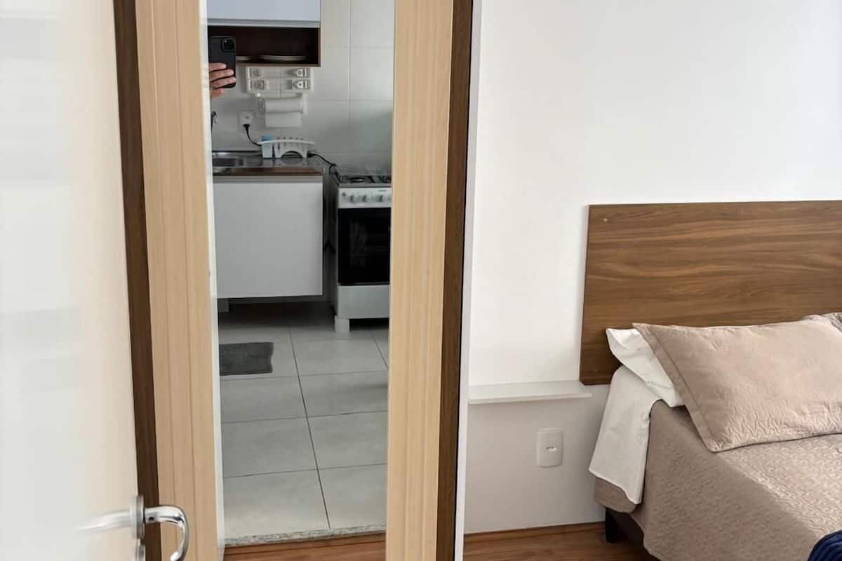 A bedroom with a wooden headboard, a bed with beige bedding, and a small kitchenette in the background.