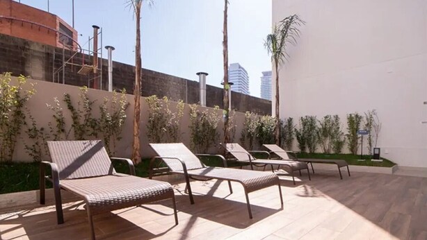 A sunlit outdoor seating area with metal chairs and a wooden floor, surrounded by plants and a white wall. - Chácara Santo Antonio - Laguna 430 (São Paulo)