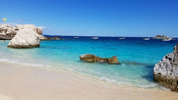 A beach with clear turquoise water, large white rocks, and a few boats in the distance. - 4½-room apartment (126m²) on ground floor with veranda and access to the garden. (Sardegna)