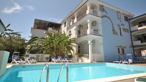 A pool area with lounge chairs, a building with balconies, and a clear sky. - 4½-room apartment (126m²) on ground floor with veranda and access to the garden. (Sardegna)