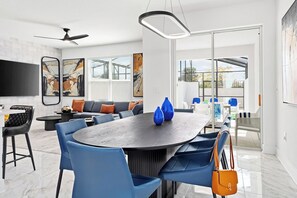 A modern dining area with a round table, blue chairs, and a sofa. There are abstract paintings on the walls and a ceiling fan. - Ivy's Isle | Mythical villa w PVT pool oasis, dragon theme BR, next to Universal (Orlando)