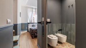 Un bagno moderno con toilette, bidet e cabina doccia in vetro. Si intravede, nella stanza adiacente, un letto con copripiumino floreale.