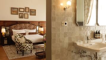 Habitación de hotel con una cama grande, un área de descanso con una silla, una mesita de noche con lámparas y un baño con lavamanos y espejo.