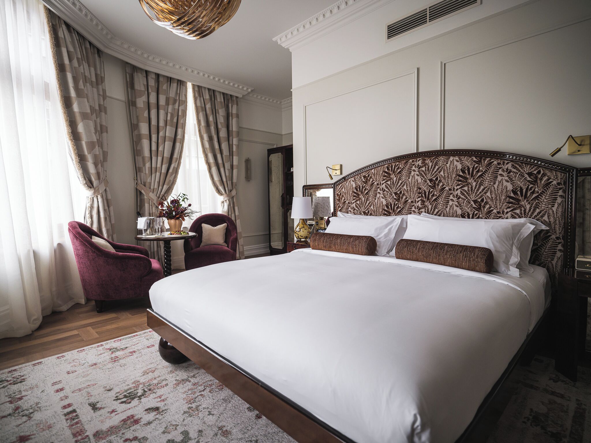 A large bed with a patterned headboard, a wooden floor, a red armchair, a small table with a vase, and a mirror on the wall.