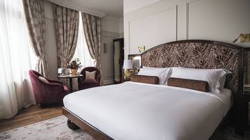A large bed with a patterned headboard, a wooden floor, a red armchair, a small table with a vase, and a mirror on the wall.