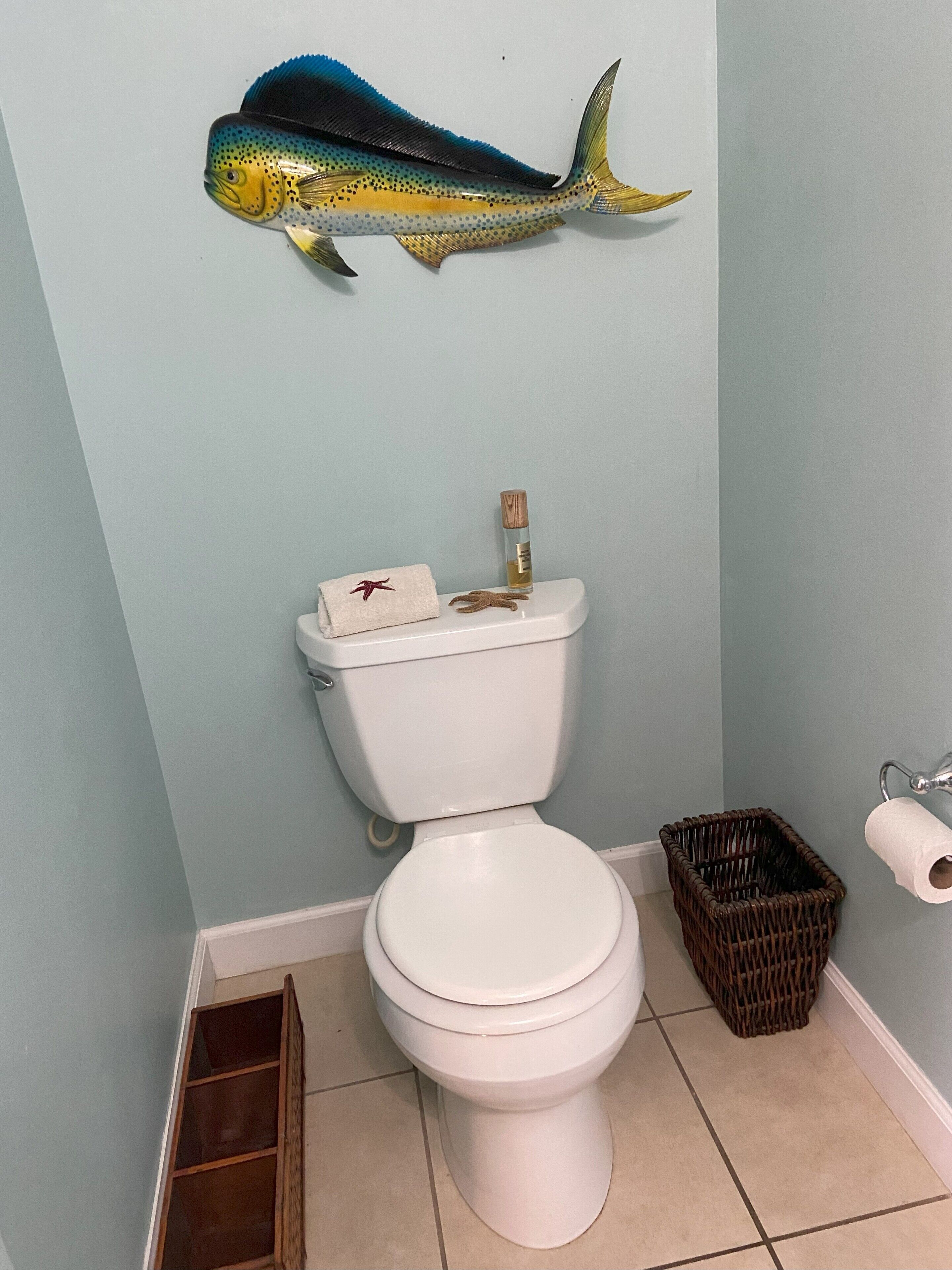 A bathroom with a toilet, a decorative fish on the wall, a bottle of hand sanitizer, a roll of toilet paper, and a wicker basket.