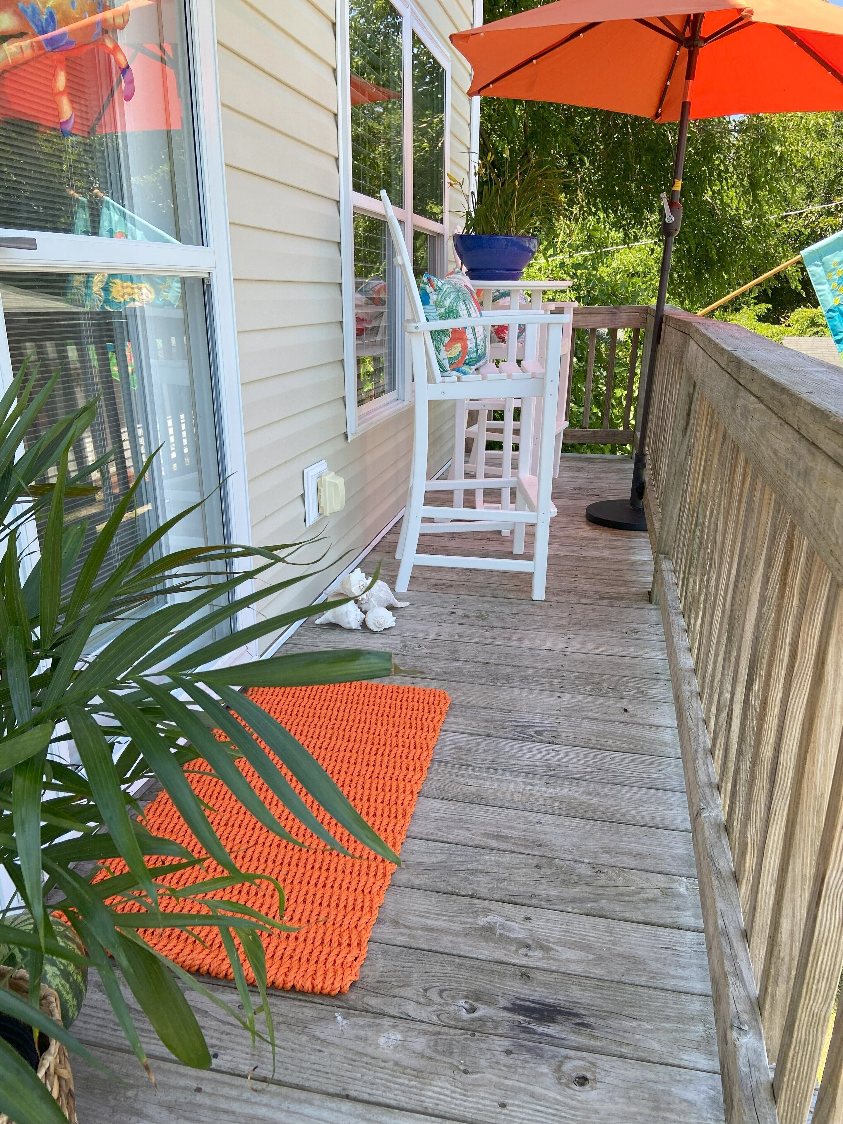 A wooden deck with a white chair and a blue pot, an orange umbrella, and a wooden railing.