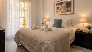 A neatly made bed with pillows and towels, a bedside table with a lamp, a framed picture on the wall, and a window with sheer curtains. - Duplex in Puerto Banus by Costarentals - Ref M40 (Marbella)