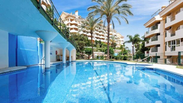 A swimming pool with a clear blue water surface, surrounded by multi-story buildings with balconies and palm trees. - Duplex in Puerto Banus by Costarentals - Ref M40 (Marbella)