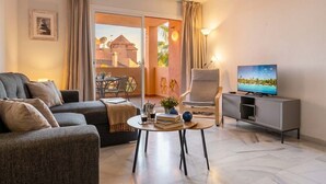 A living room with a sofa, a coffee table with books and a plant, a TV on a stand, and a view of a building outside. - Reserva De Marbella apt by Costarentals - Ref M43 (Artola)