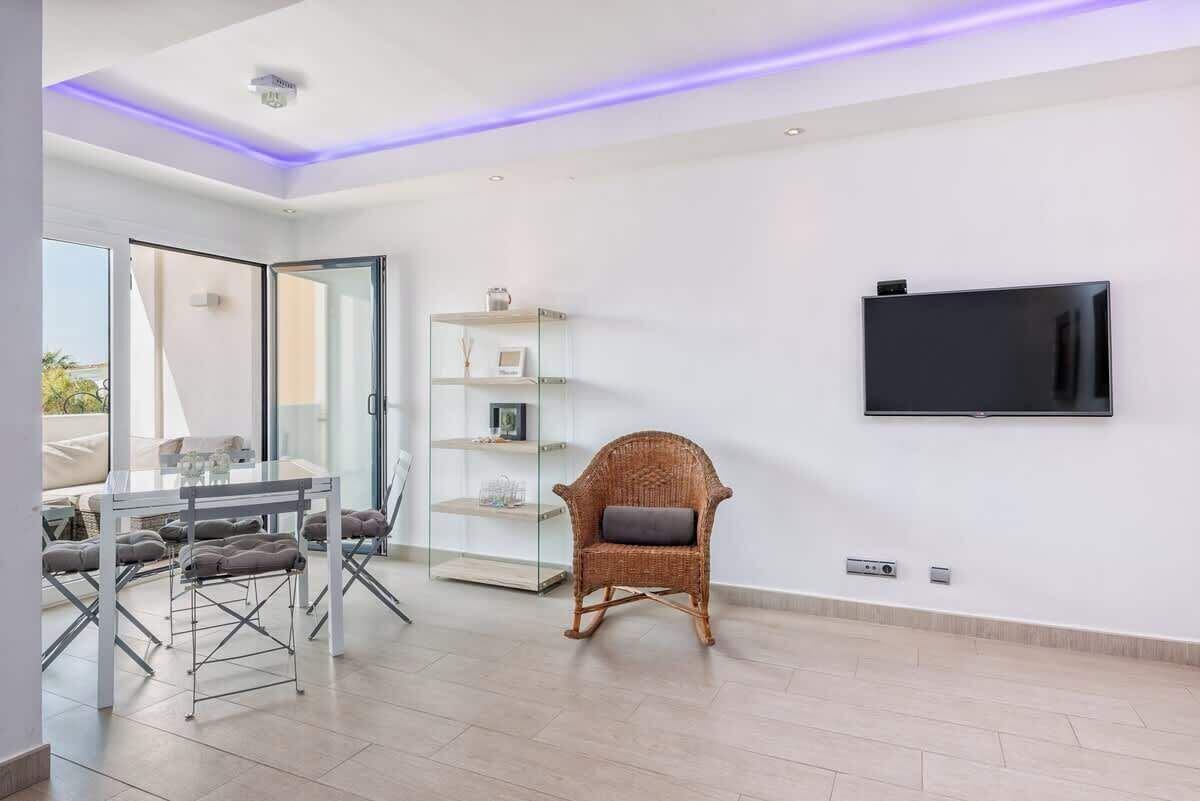 Een moderne woonkamer met een glazen eettafel, een rieten stoel en een tv aan de muur.