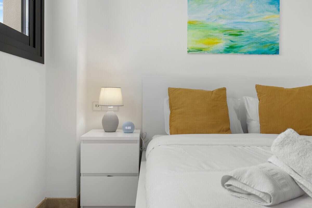 Une chambre à coucher bien rangée, avec un lit, des oreillers, une table de chevet avec une lampe et une horloge murale.
