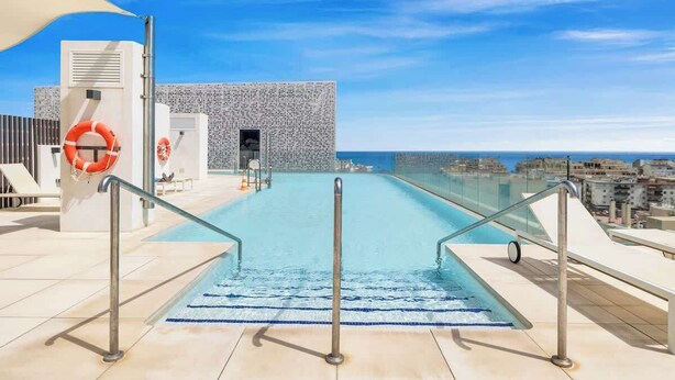 A rooftop pool with a clear view of the city skyline, a lifebuoy, and a glass railing.