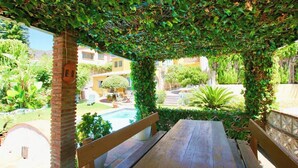 A wooden bench under a pergola with climbing vines, overlooking a swimming pool and a garden.
