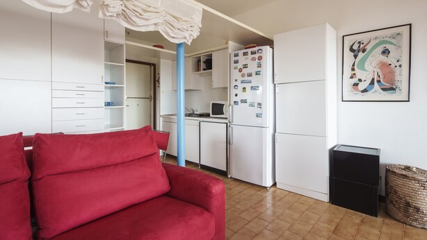 Un espacio habitable compacto con kitchenette, un sofá rojo y un cuadro abstracto enmarcado.