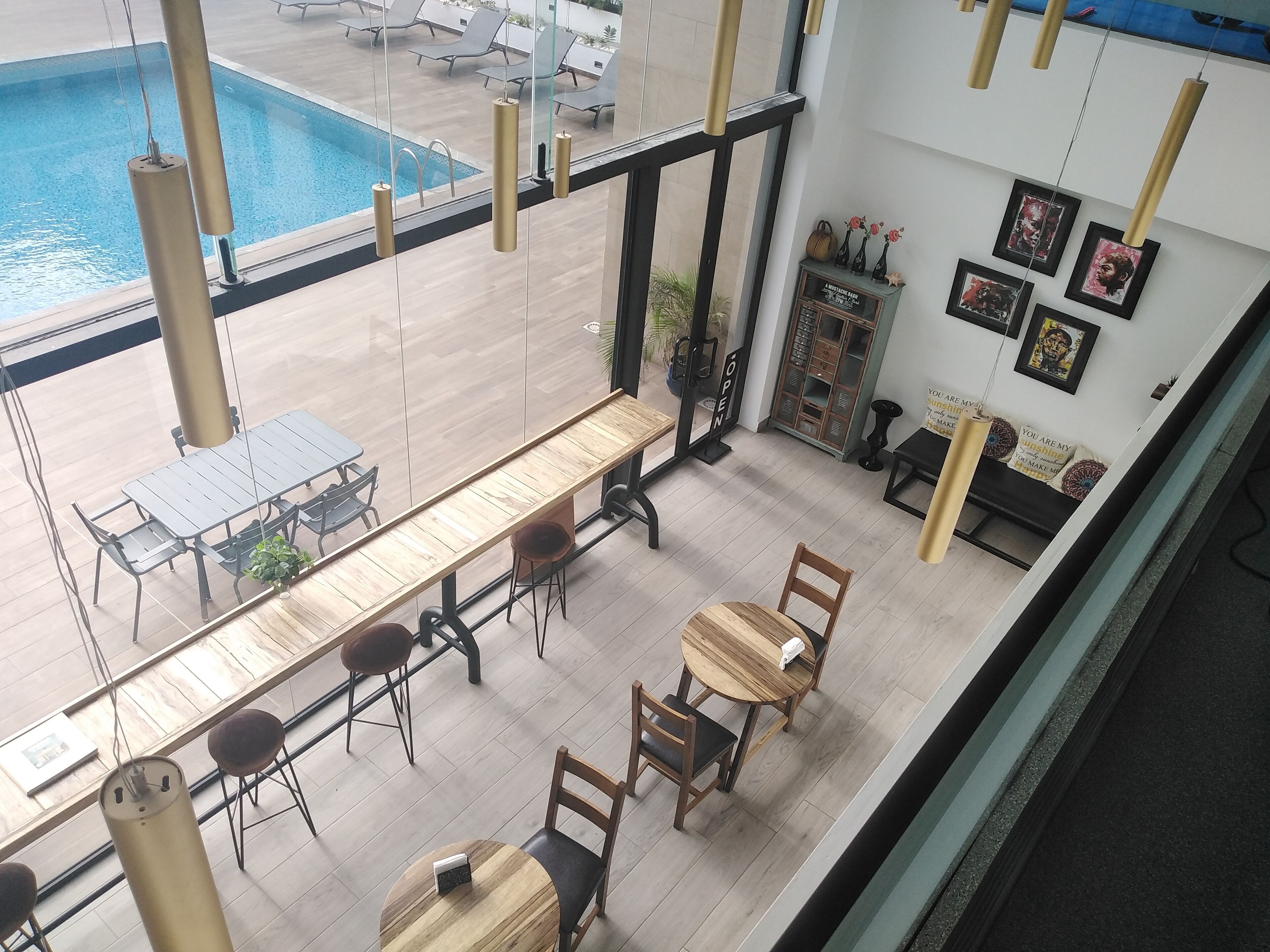 Un espace aménagé au bord de la piscine, avec une longue table et des chaises en bois, un petit coin salon avec un banc et des chaises, et un mur orné de tableaux et sur lequel est installé un meuble.